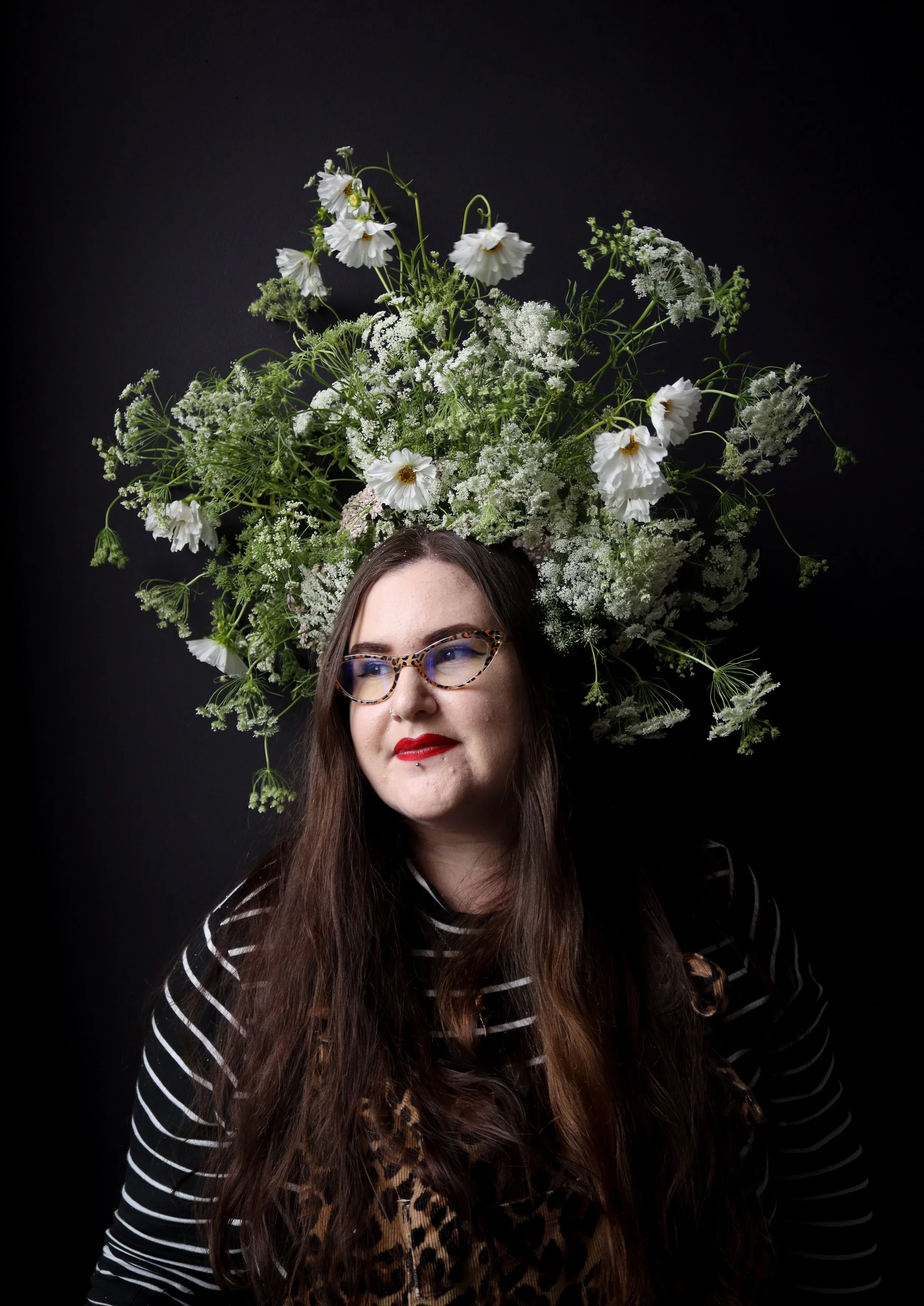 Sarah has long brown hair, is wearing glasses with leopard print frames and red lipstick. She's looking slightly to the side against a black background. She has a large arrangement of white flowers and greenery on her head, resembling a crown.