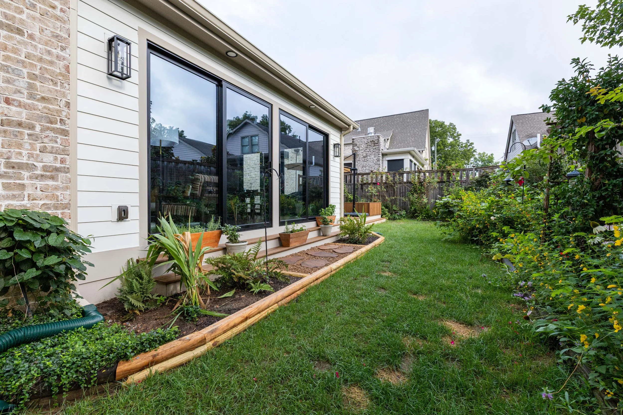 Backyard garden with plants, flowers, a wooden border, and a house with large glass sliding doors.