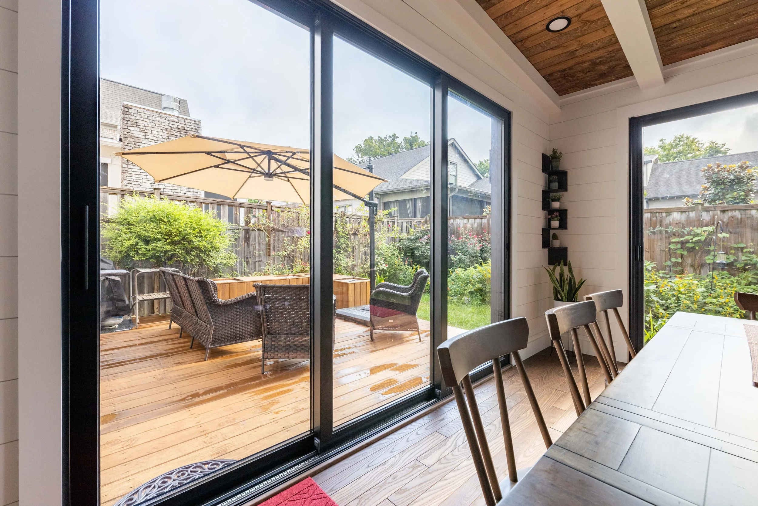 View from inside a house looking out through glass sliding doors onto a wooden deck with outdoor furniture, an umbrella, and a garden with plants, trees, and neighboring houses in the background.