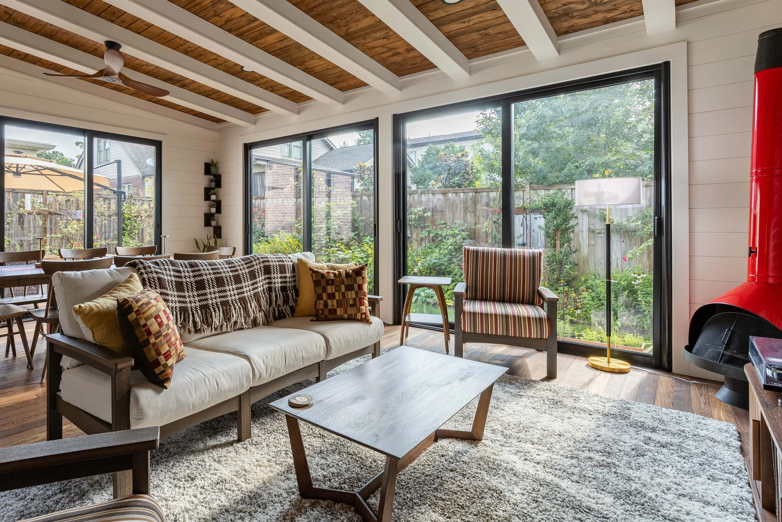 Living room with large glass sliding doors showing a backyard garden, white walls, a wooden ceiling with exposed beams, a ceiling fan, a beige couch with patterned pillows, a wooden coffee table, a striped armchair, a tall white lamp, and a red wood 