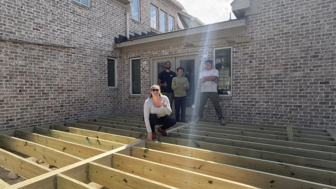 Four people, two women and two men, standing and kneeling on a wooden deck frame under construction outside a brick house on a sunny day.
