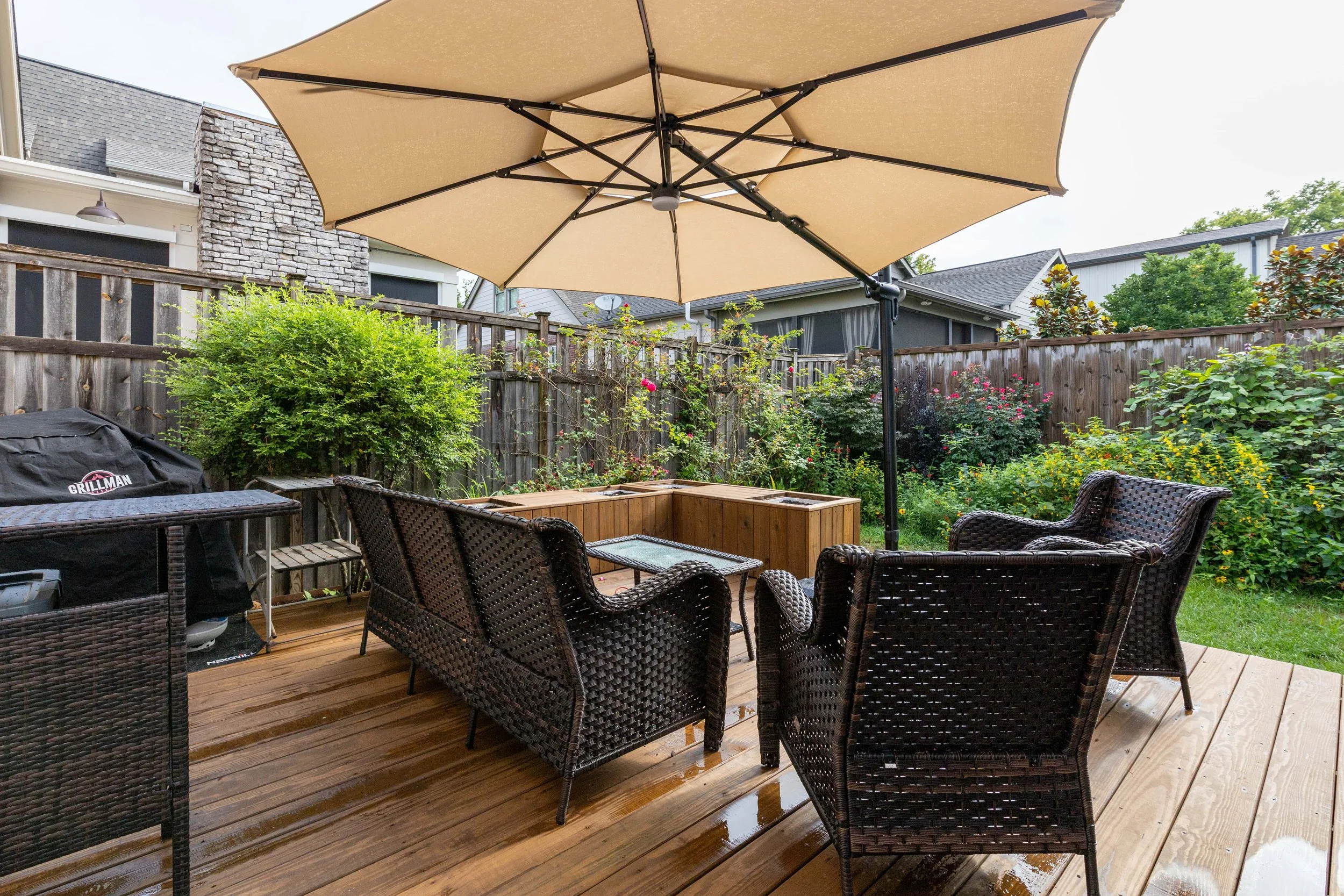 Backyard patio with wicker furniture including chairs and a glass-topped table, a large beige umbrella providing shade, a wooden deck, surrounding wooden fence, lush green plants, flowering bushes, a grill, and neighboring houses with a cloudy sky in