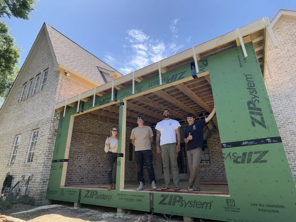 Four workers standing on the porch of a house under construction, with green ZIP System sheathing, a brick exterior, and a partially built roof. The workers are smiling and posing for the photo.