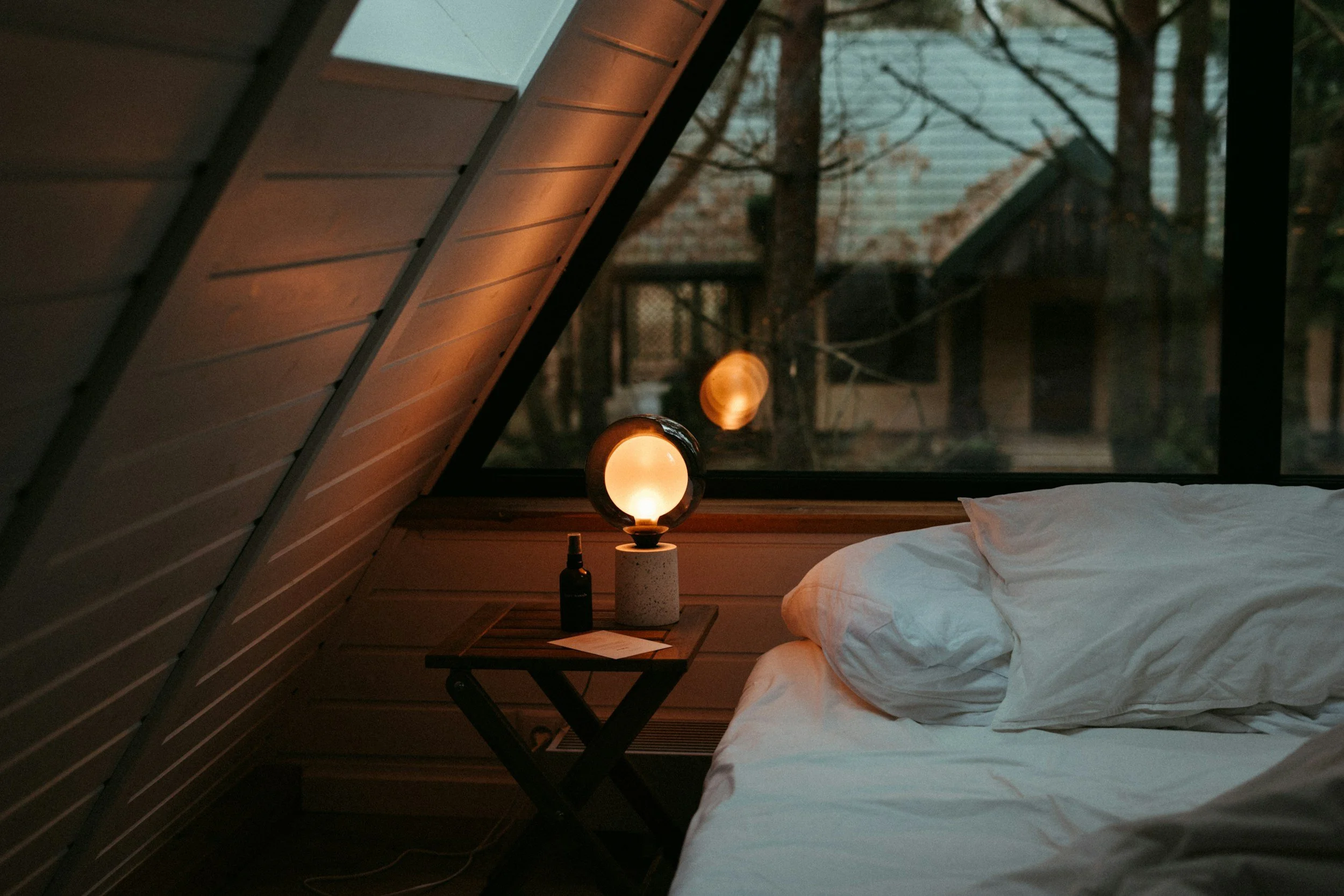 A cozy bedroom corner with a bed that has white linens, a small nightstand with a lamp, a sketchbook, and a small bottle. A large window shows a view of trees outside, indicating a natural, wooded setting.