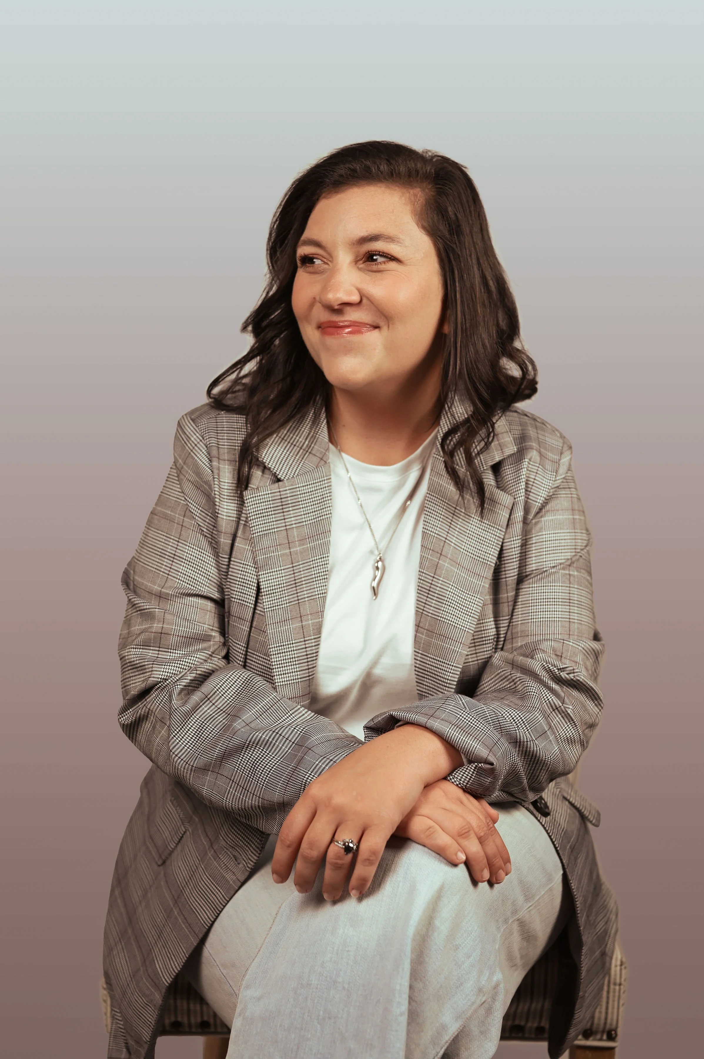 A woman with dark hair wearing a checked blazer and white shirt, sitting with crossed arms, smiling and looking to her left against a gradient background.