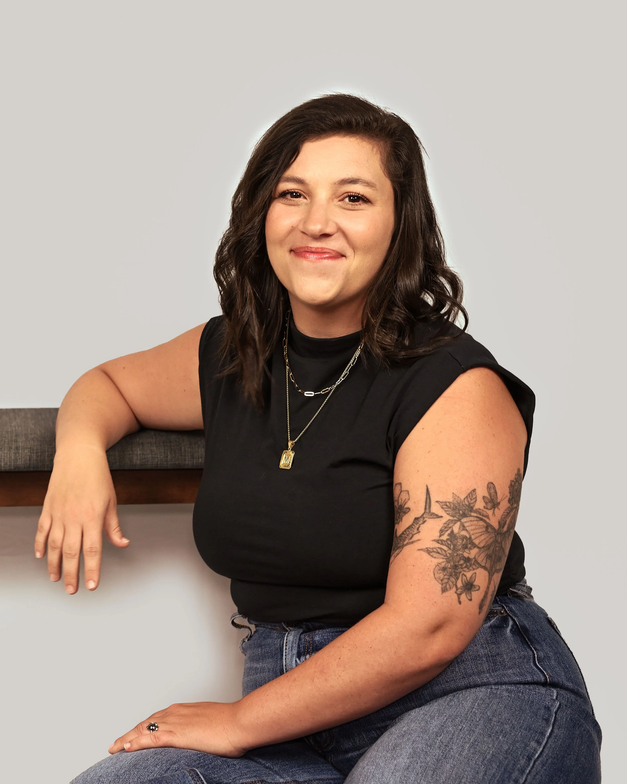 A young woman with long, wavy brown hair and tattoos on her left arm, wearing a black sleeveless top and jeans, sitting against a plain background with a neutral expression.