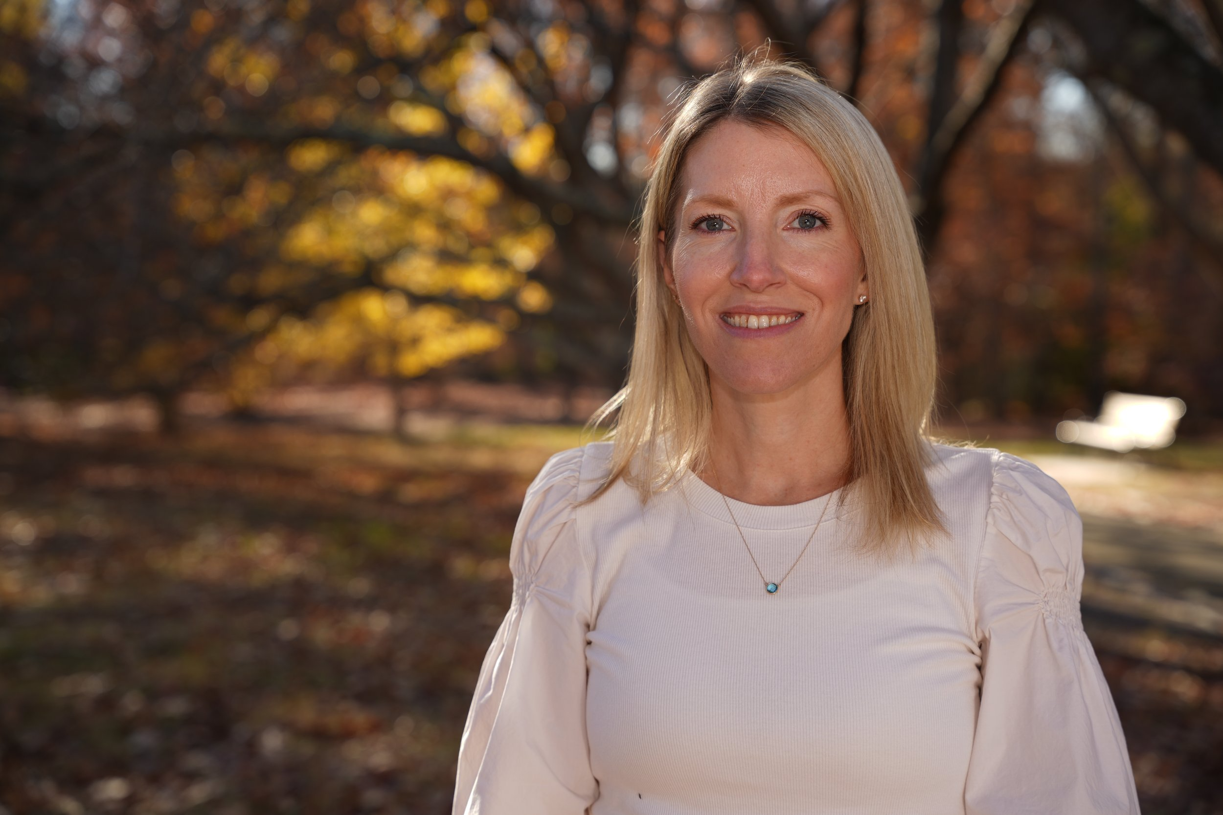 A smiling woman with blonde hair wearing a white top and a necklace, outdoors in an autumn setting with trees and golden leaves in the background.