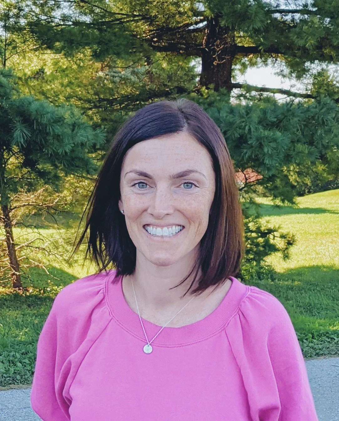 A woman with shoulder-length dark brown hair and blue eyes smiling outdoors in front of green trees, wearing a pink top and a silver necklace.