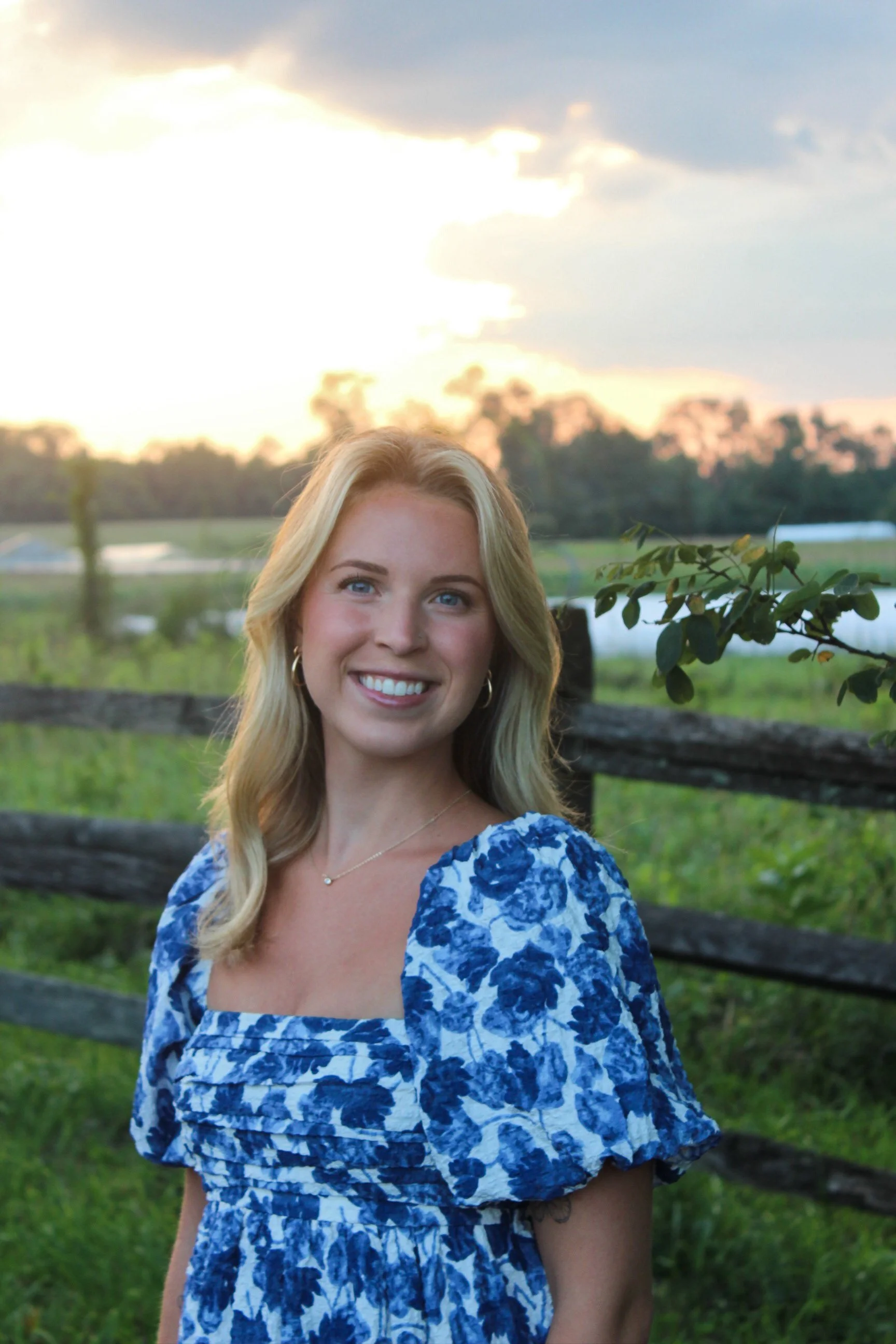 A smiling woman with blonde hair, wearing a blue and white floral dress, outdoors near a river or pond, with a sunset sky and trees in the background.