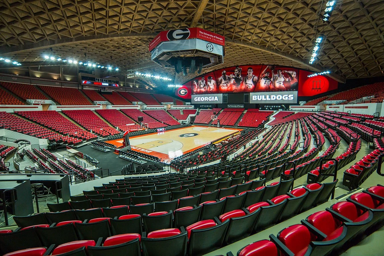 Empty basketball arena with red seats, scoreboard displaying Georgia and Bulldogs, and a large screen showing basketball players.