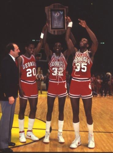 Three basketball players in red jerseys celebrating on a basketball court, holding up a large trophy. The player in the middle is wearing jersey number 32 and the other two are wearing numbers 20 and 35. A man in a suit is standing to the left, smiling.