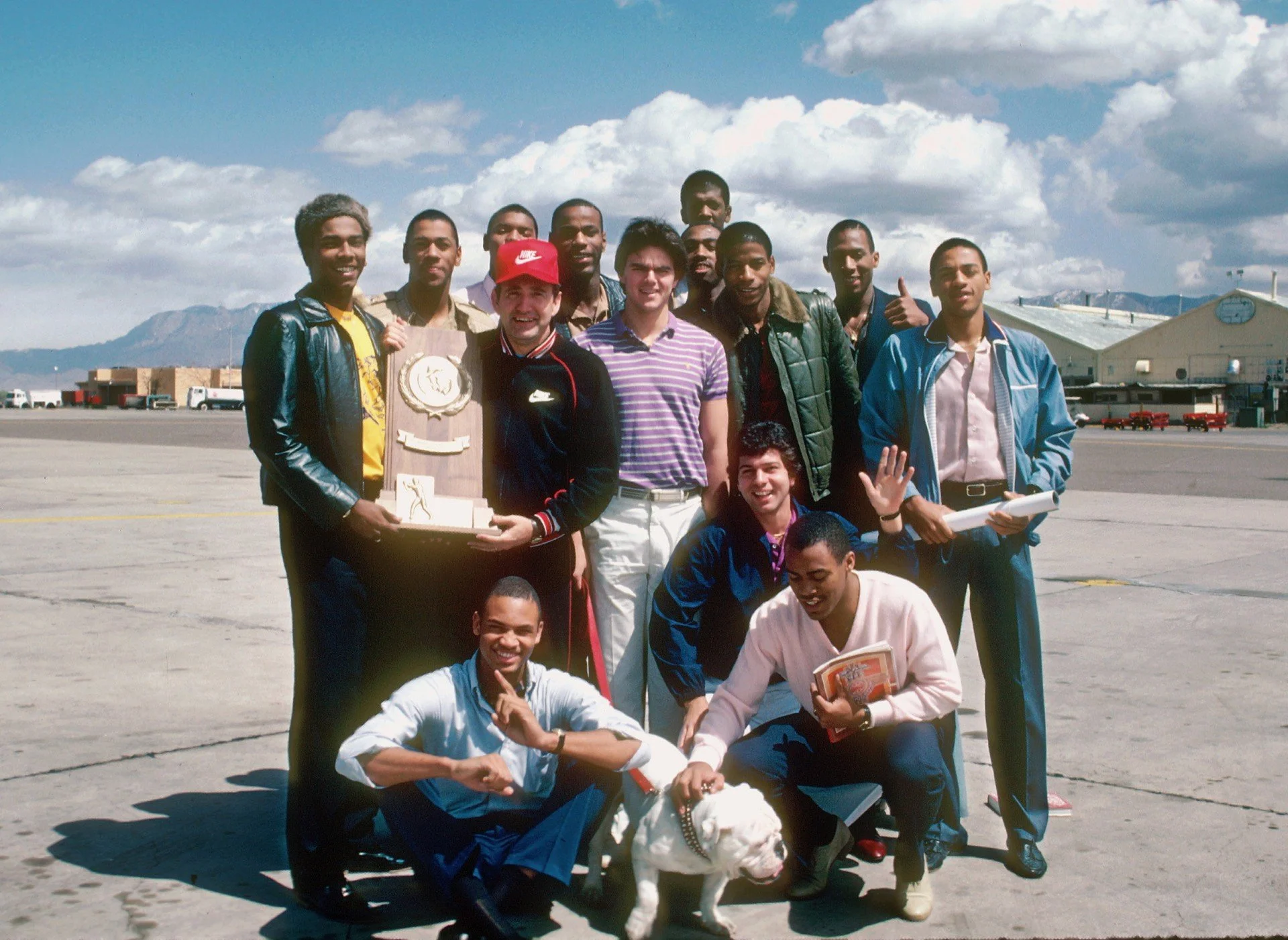 Group of men and boys posing outdoors at an airport with mountains and blue sky in the background. One man is holding a trophy. Some are smiling or making gestures, and one is holding a dog. The group appears to be celebrating or commemorating an event.