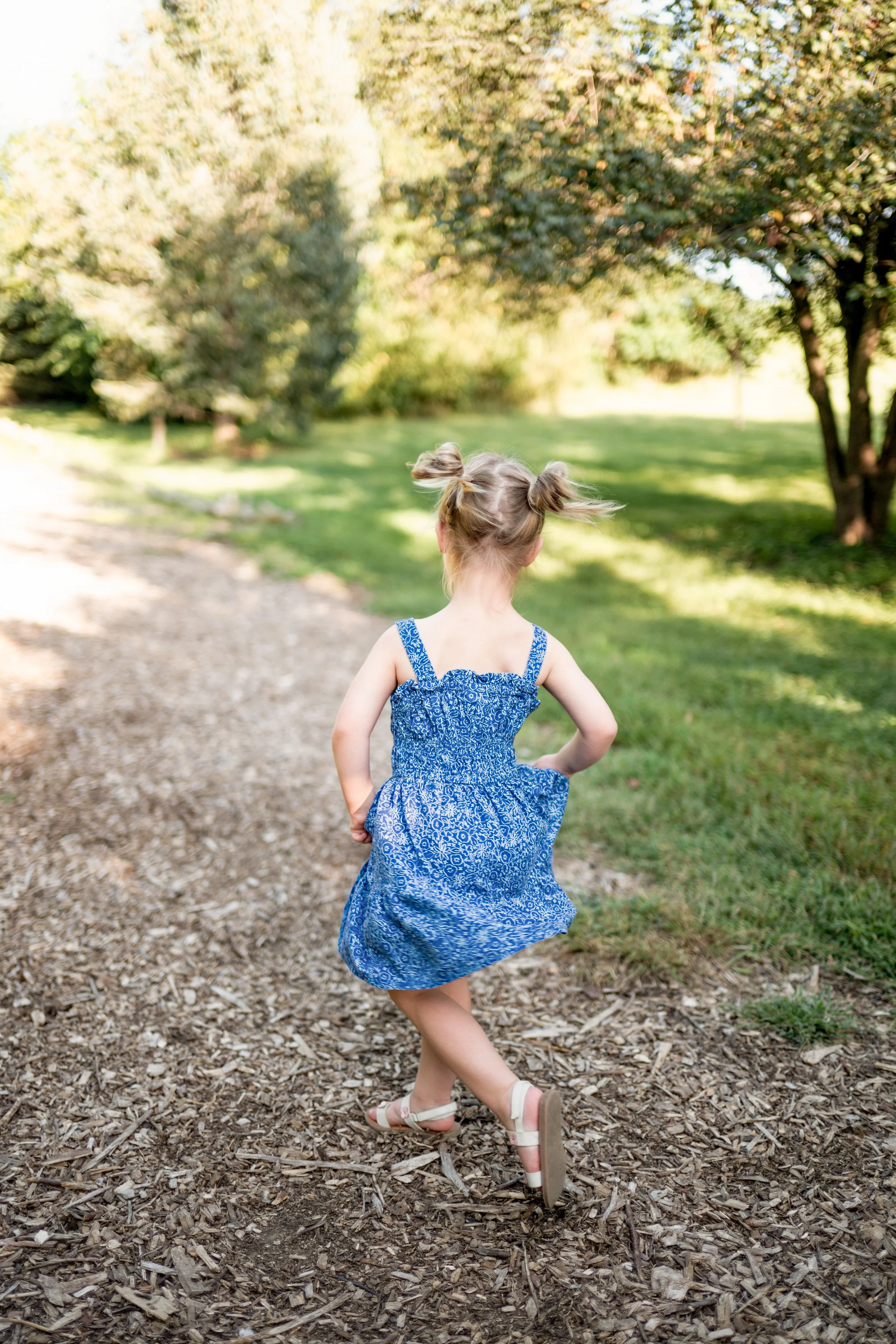 A young girl with blonde hair styled in two buns, wearing a blue patterned dress and sandals, is running on a dirt path in a park with trees and green grass.