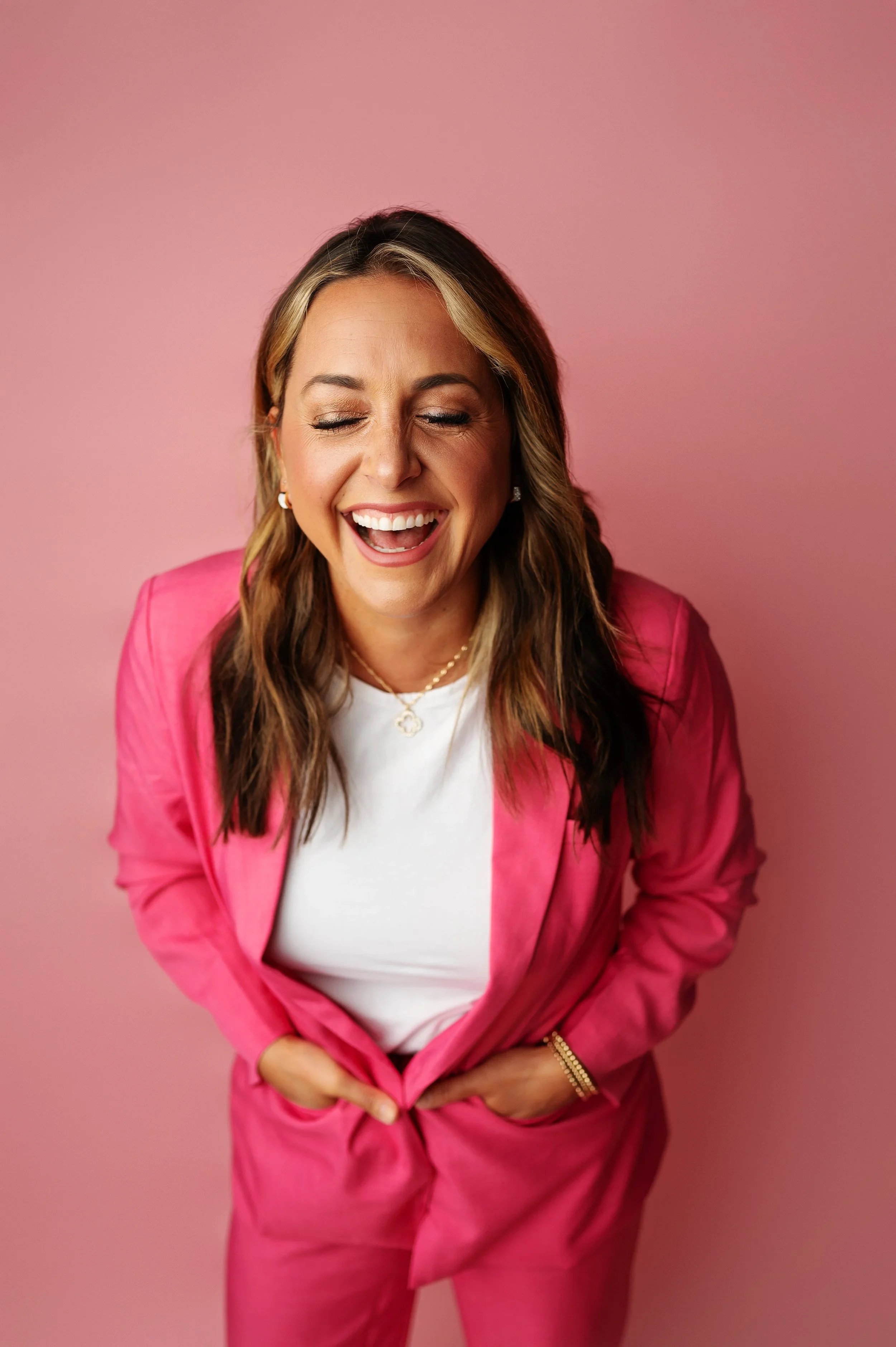 A woman laughing with her eyes closed, wearing a pink blazer, white t-shirt, and jewelry, standing against a pink background.