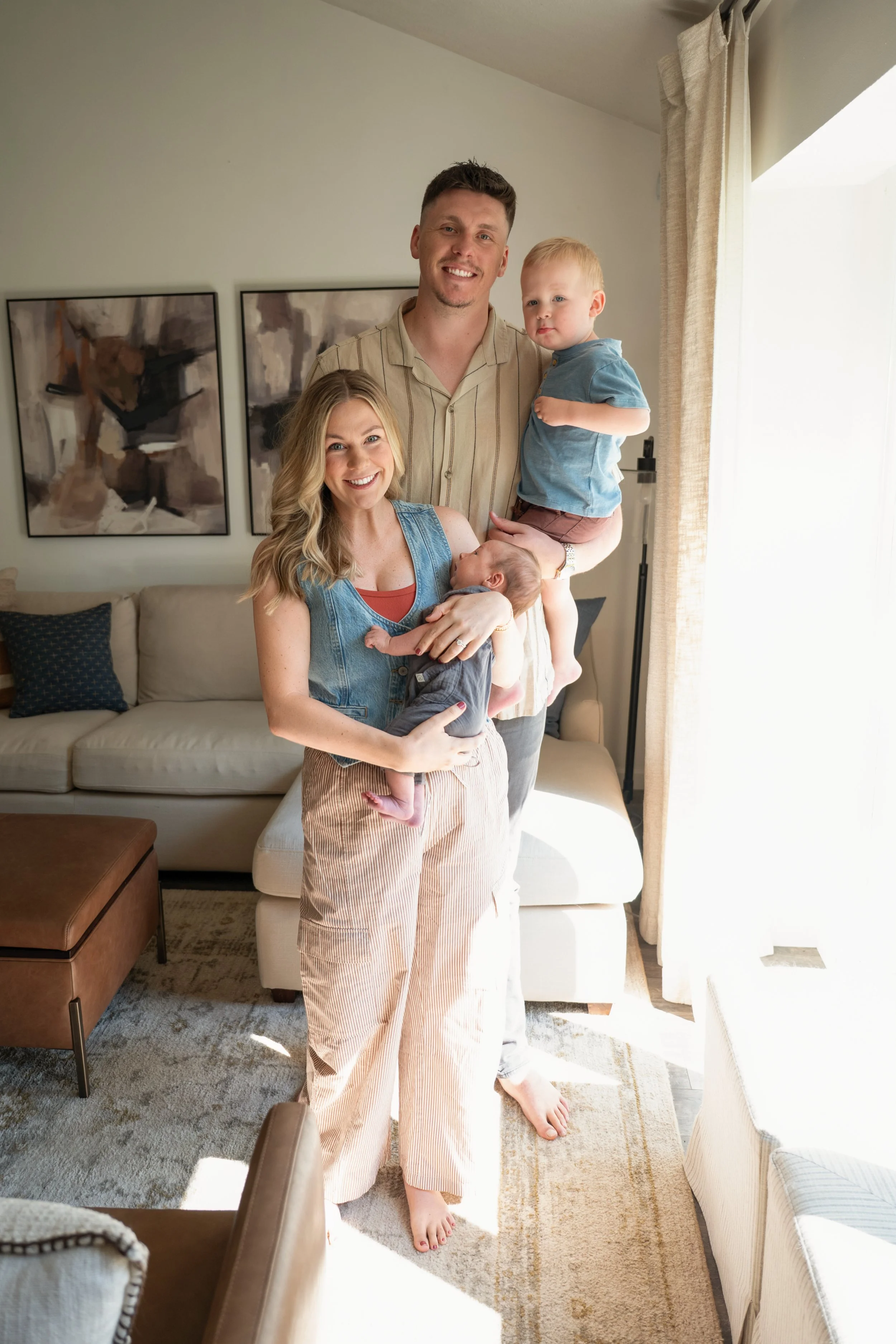 A family of four in a living room. A woman is holding a baby, a man is holding a young boy, all smiling. The room has a beige sofa, patterned pillows, and abstract artwork on the wall. Bright sunlight enters through curtains.