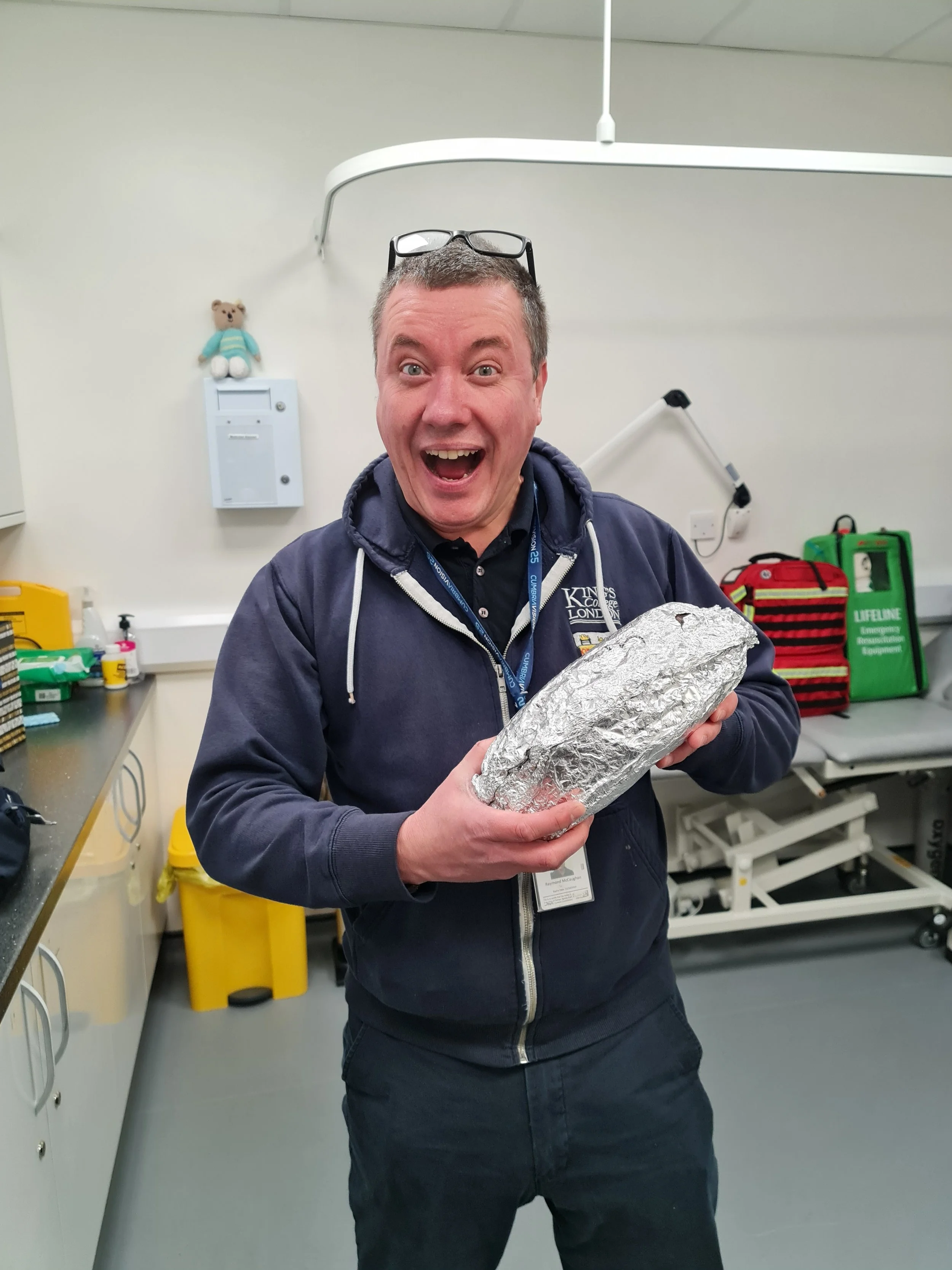 A man smiling and holding a foil-wrapped item in a medical or laboratory setting with medical supplies and equipment in the background.