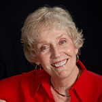 Smiling elderly woman with curly gray hair wearing a red top against a black background.