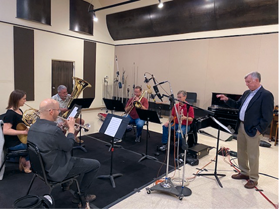 A group of musicians practicing in a rehearsal room, with a conductor standing nearby.