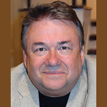 Close-up of a smiling middle-aged man with short dark hair and a light-colored blazer.