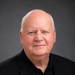 Close-up portrait of an elderly man with a serious expression, wearing a black suit jacket, against a dark grey background.