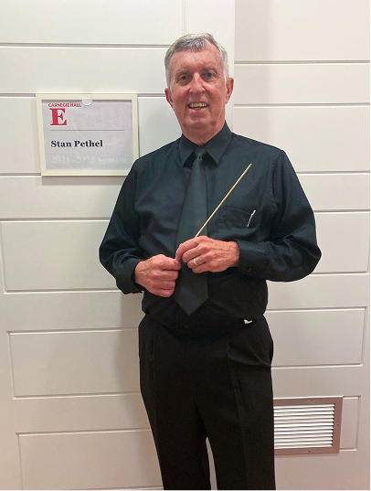 Man in black dress shirt and tie holding a conductor's baton standing in front of a wall with a sign reading 'Stan Phelan' at Carnegie Hall.