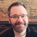 Close-up portrait of a man with glasses, smiling, with a brick wall background.