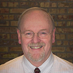 Portrait of a smiling, middle-aged man with glasses, short light-colored hair, and a beard, wearing a white shirt and a tie, standing in front of a brick wall.