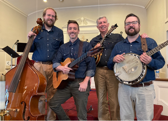 Group of four men standing together in a room, holding musical instruments including a double bass, an acoustic guitar, a ukulele, and a banjo.