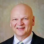 Close-up portrait of a bald man in a suit and tie smiling at the camera.