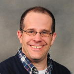 A smiling man wearing glasses, a checkered shirt, and a dark sweater.