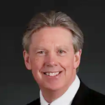 A smiling middle-aged man with gray hair, wearing a dark suit and white shirt, against a dark background.