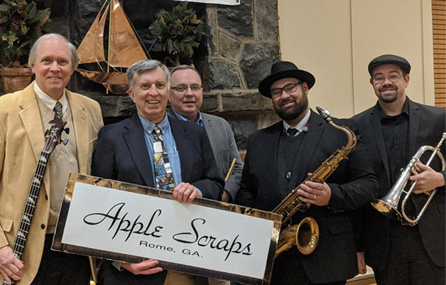 Five men standing indoors, holding musical instruments and a sign that says 'Apple Scraps Rome, GA.' The men are dressed in semi-formal attire, and they are smiling.