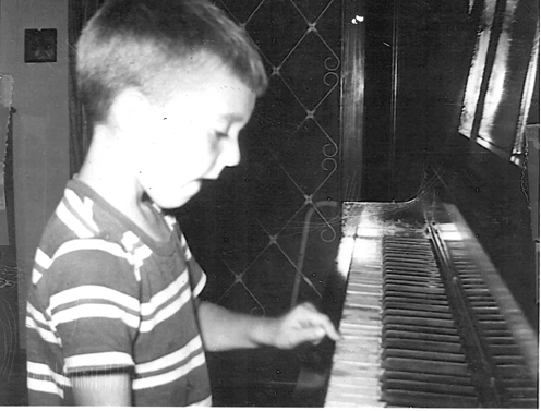 A young boy in a striped shirt looking at a piano in a room.
