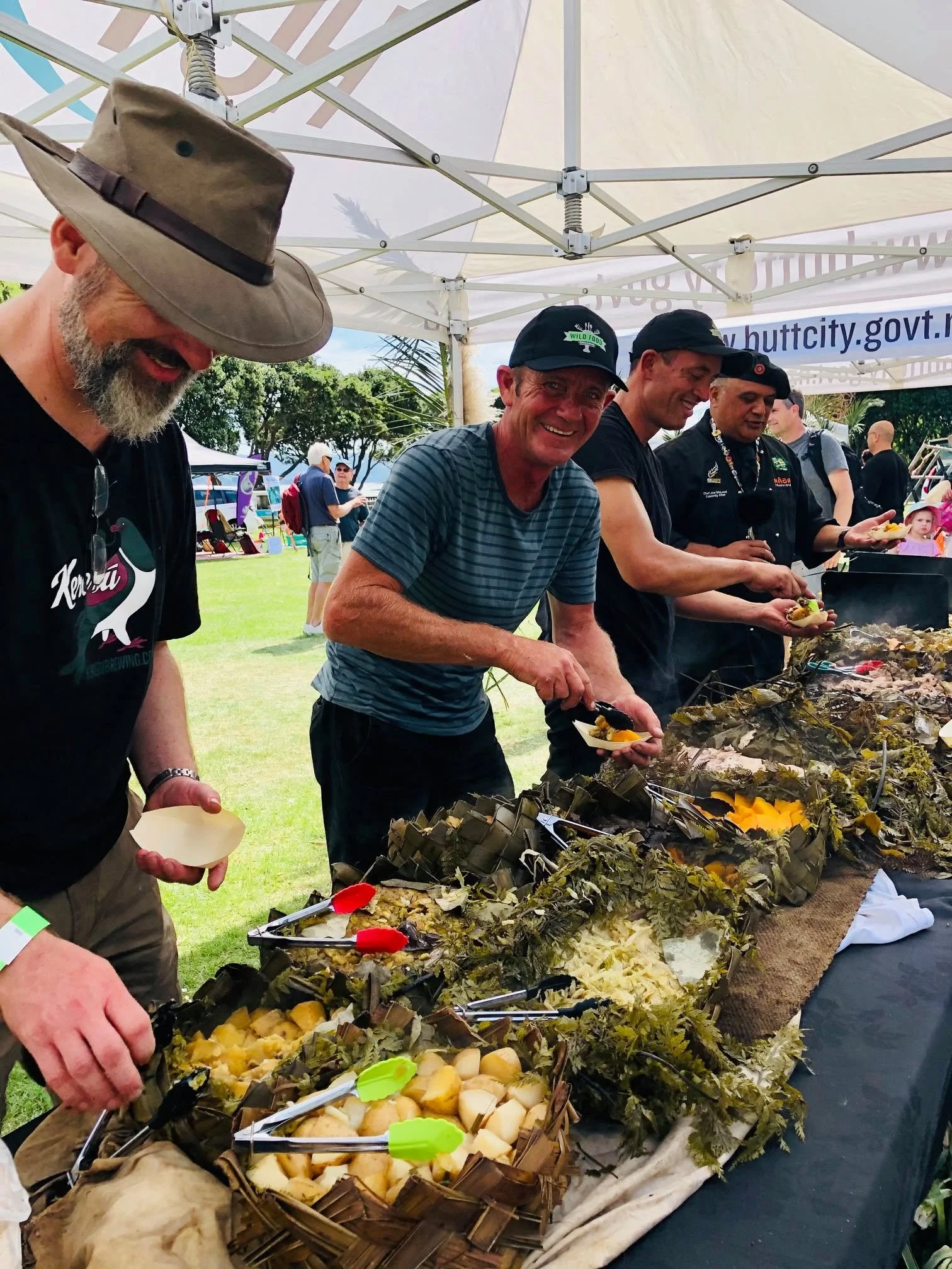 People gathered under a white canopy at an outdoor event, serving and eating food from a long table decorated with leaves and flowers.