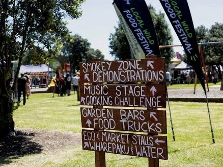 Signboard with directions at a local food festival, pointing to smoky zone, music stage, garden bars, food trucks, eco and market stalls, wharepaku, and water, in a park area with tents and people in the background.