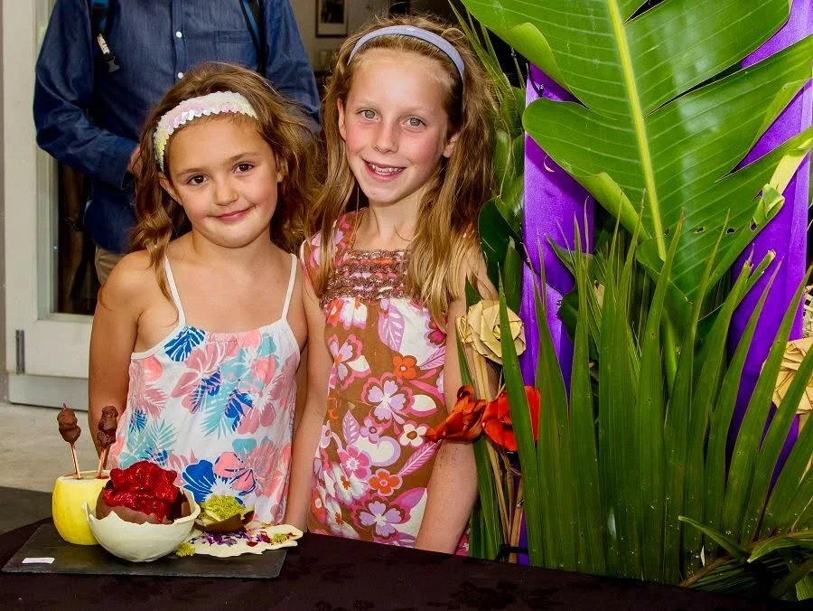 Two young girls smiling at a party, standing behind a table with decorated desserts and surrounded by green plants and purple decorations.