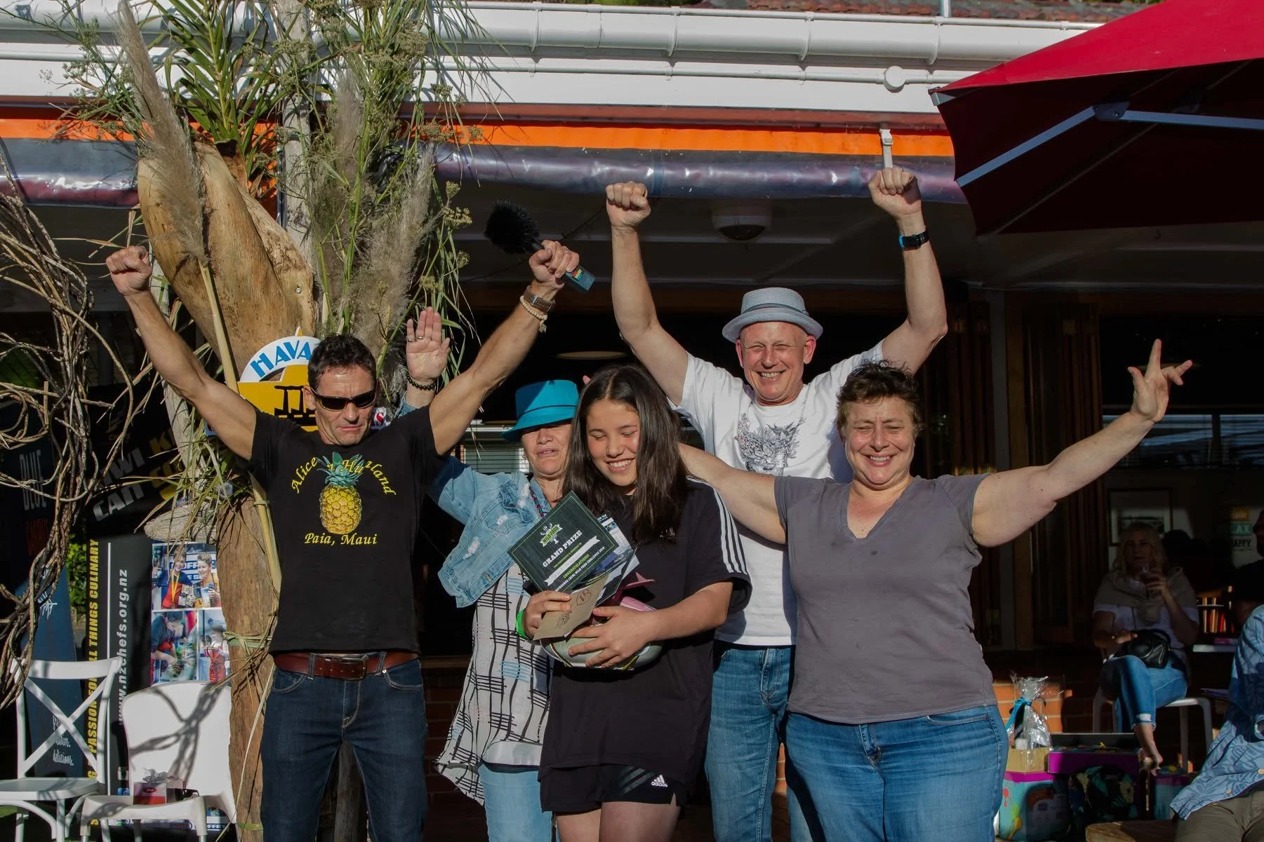Group of five people celebrating outdoors, smiling, with arms raised, holding certificates and a microphone, near a tree and umbrellas.