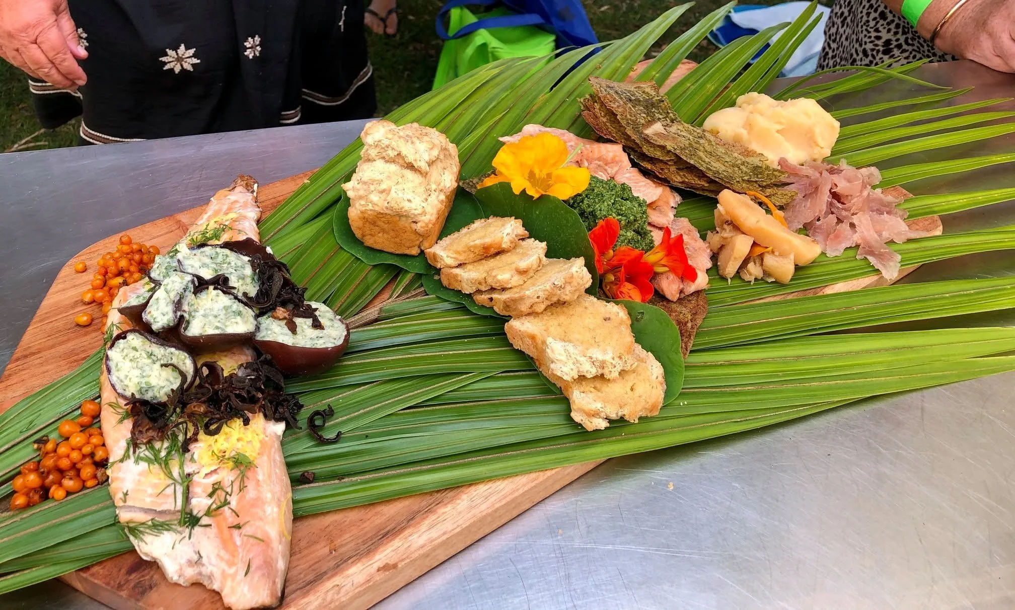 A wooden platter with various Hawaiian-style foods on large green palm leaves, including slices of bread, fresh vegetables, and salads, at an outdoor event.