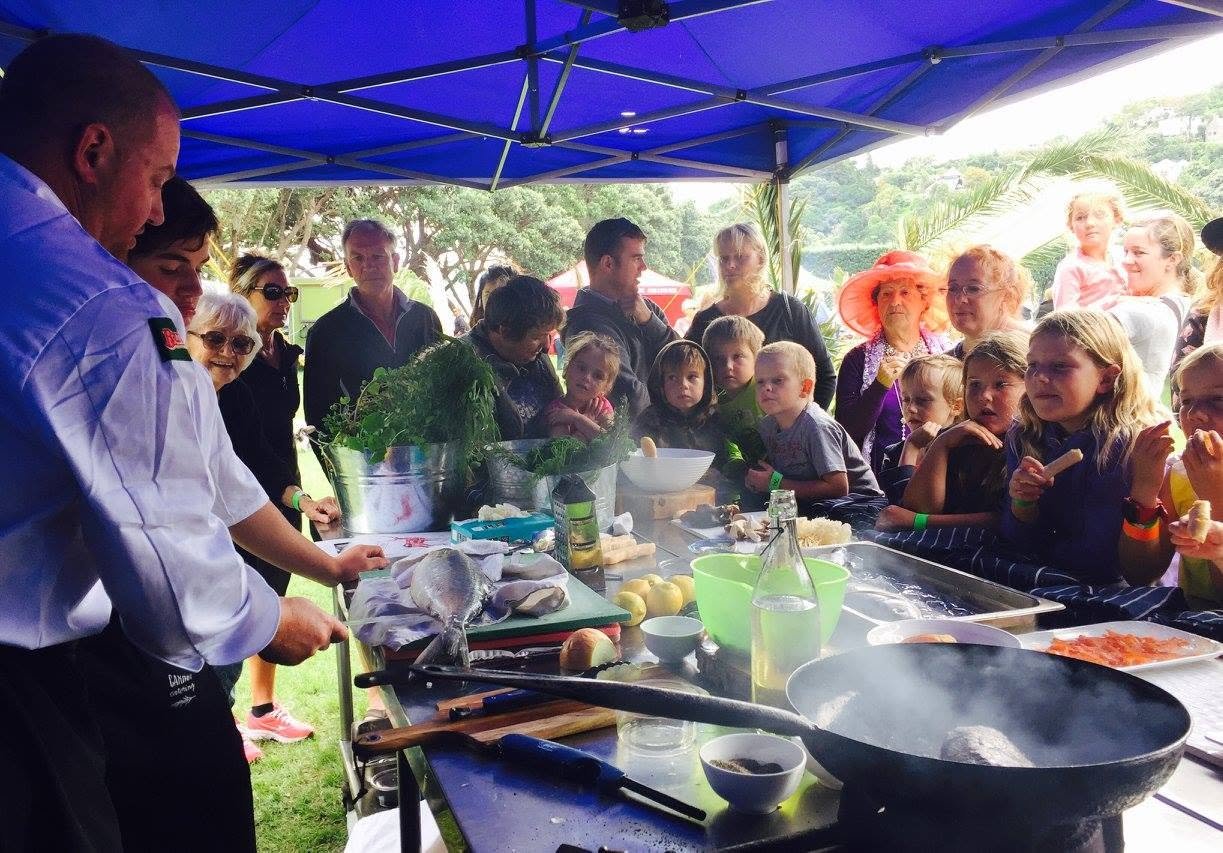 Chef demonstrating cooking techniques on an outdoor stage to a group of children and adults under a blue canopy, with cooking ingredients and utensils on the table.