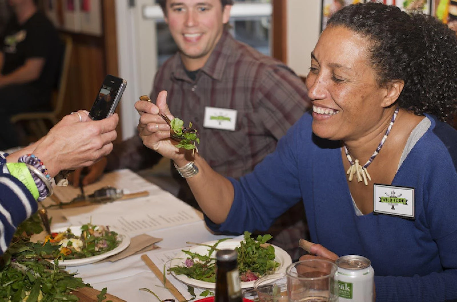People sitting at a table with salads, one woman handing a salad to another woman who is smiling, and a man smiling in the background.