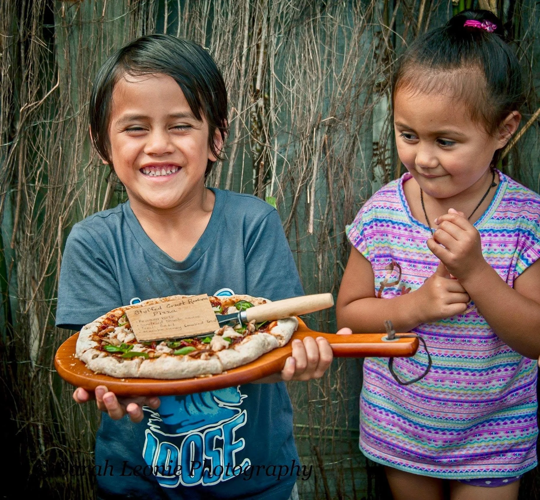 Two children, a boy and a girl, standing outdoors with a backdrop of tall grass or reeds. The boy is smiling and holding a plate with a pizza, while the girl is looking at the pizza with her hands clasped together.