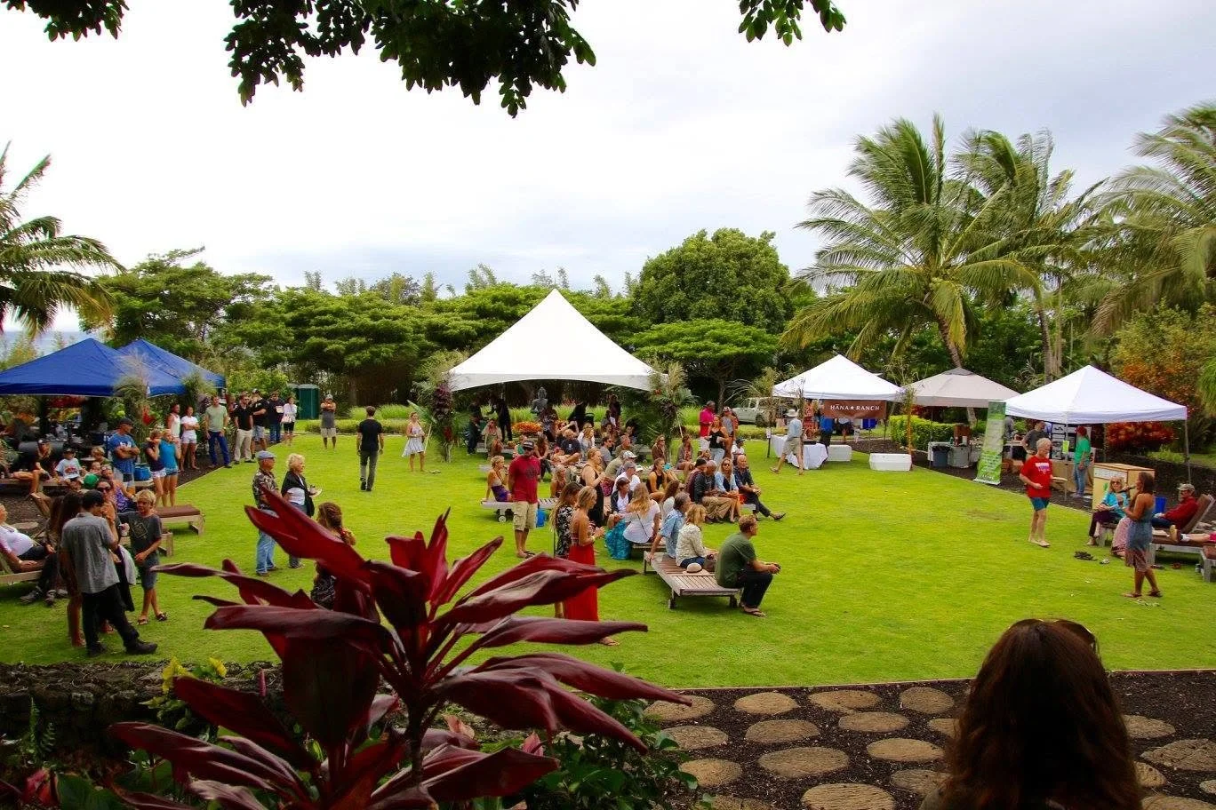 An outdoor gathering with people seated on benches on a grassy lawn, under tents, with trees including palm trees in the background and a cloudy sky.