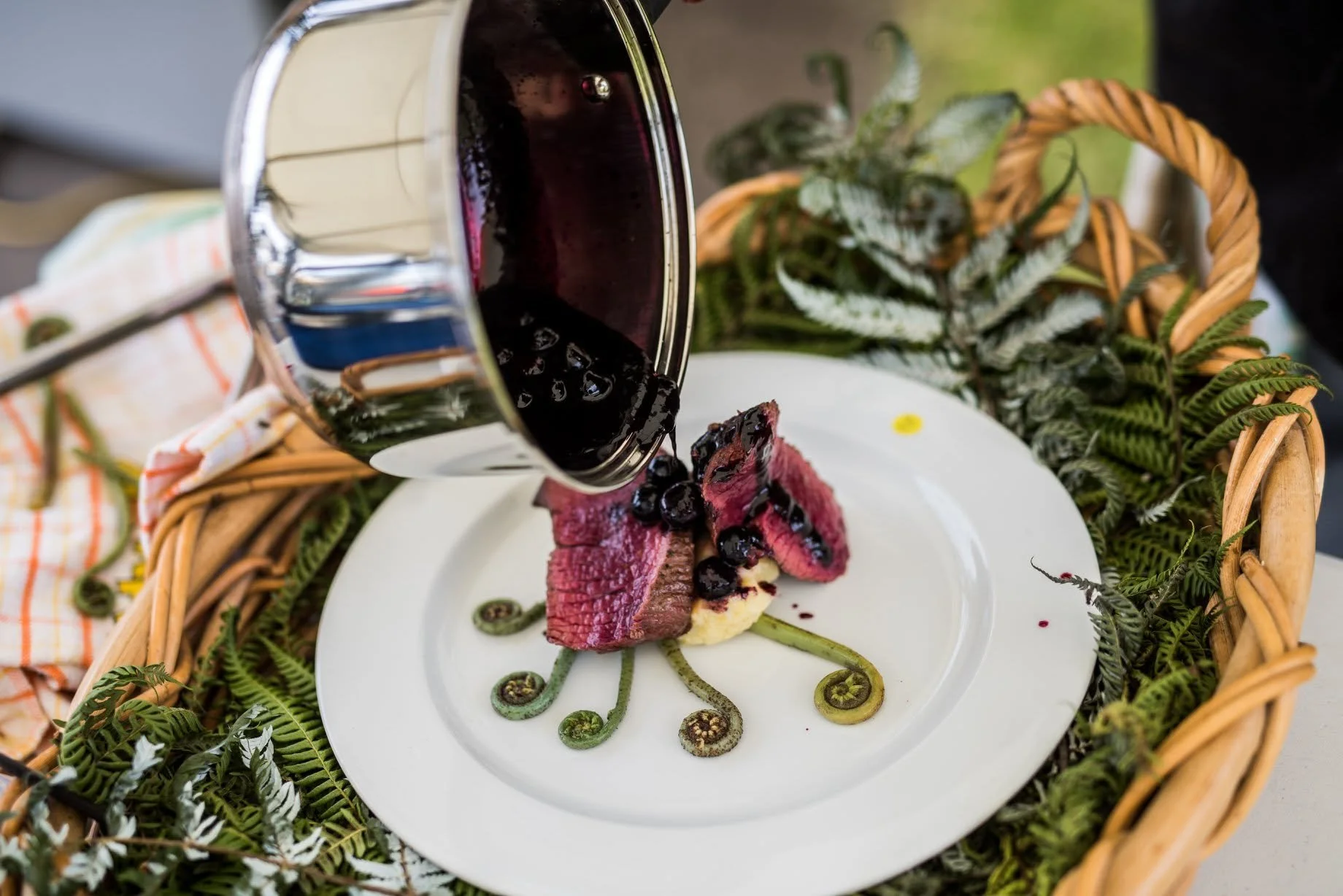 A white plate with a Charlotte Russe with a rose-like design and a stalk of green fern-shaped decoration underneath. A piece of cooked beef with a berry sauce is being poured over it from a pitcher. The dish is placed in a basket with fern leaves and