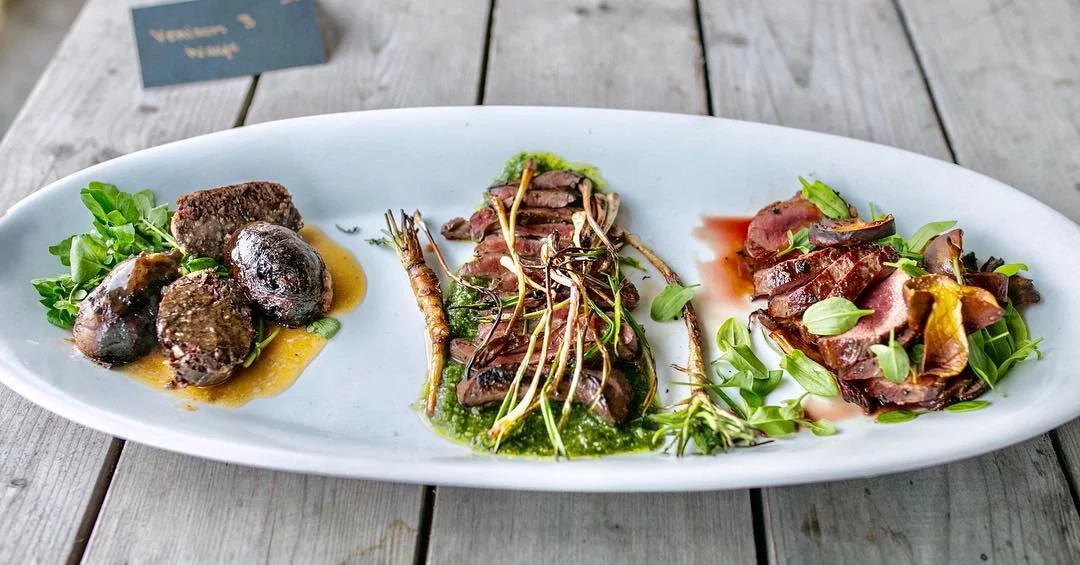 A long white oval plate with three different types of gourmet beef appetizers, garnished with herbs and sauces, on a wooden table.