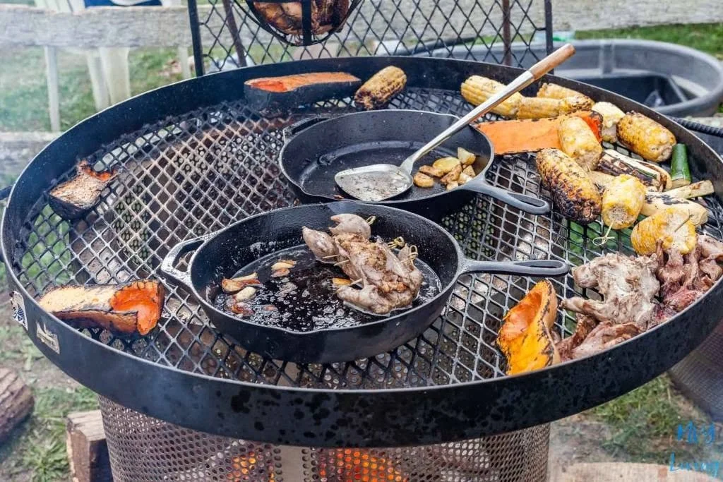 Charcoal grill with assorted vegetables and meats, including corn on the cob, sweet potatoes, chicken, and beef, cooking outdoors.