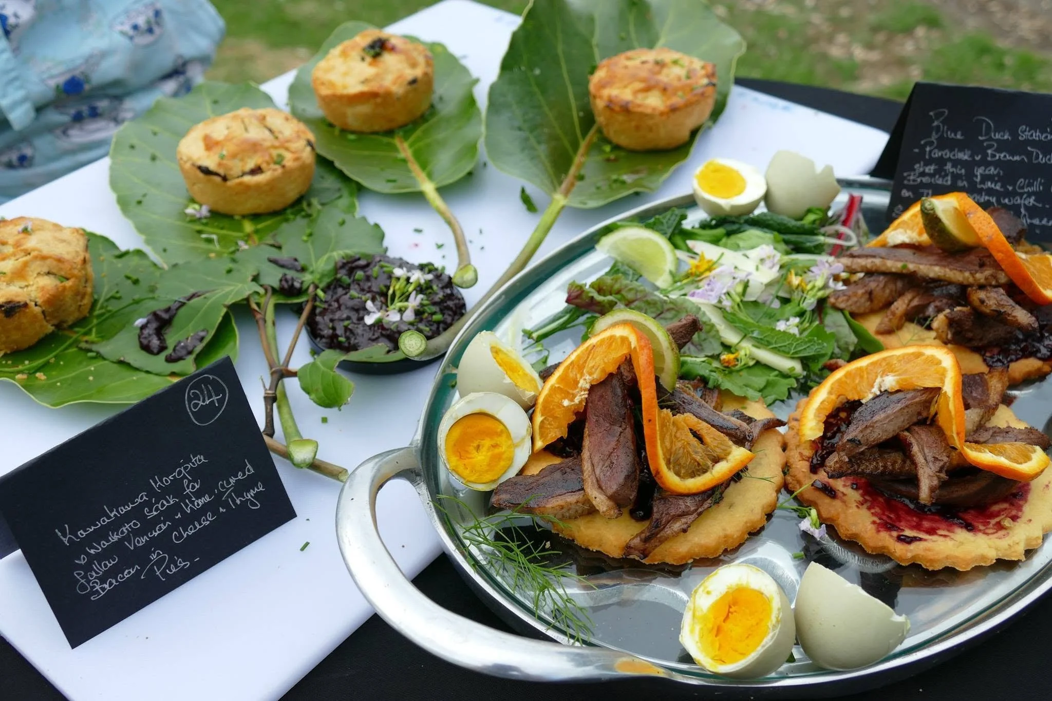 Close-up of a food display featuring baked cupcakes topped with herbs, lemon, and other garnishes, along with a salad with sliced oranges and lemon wedges, and handwritten labels describing the dishes.