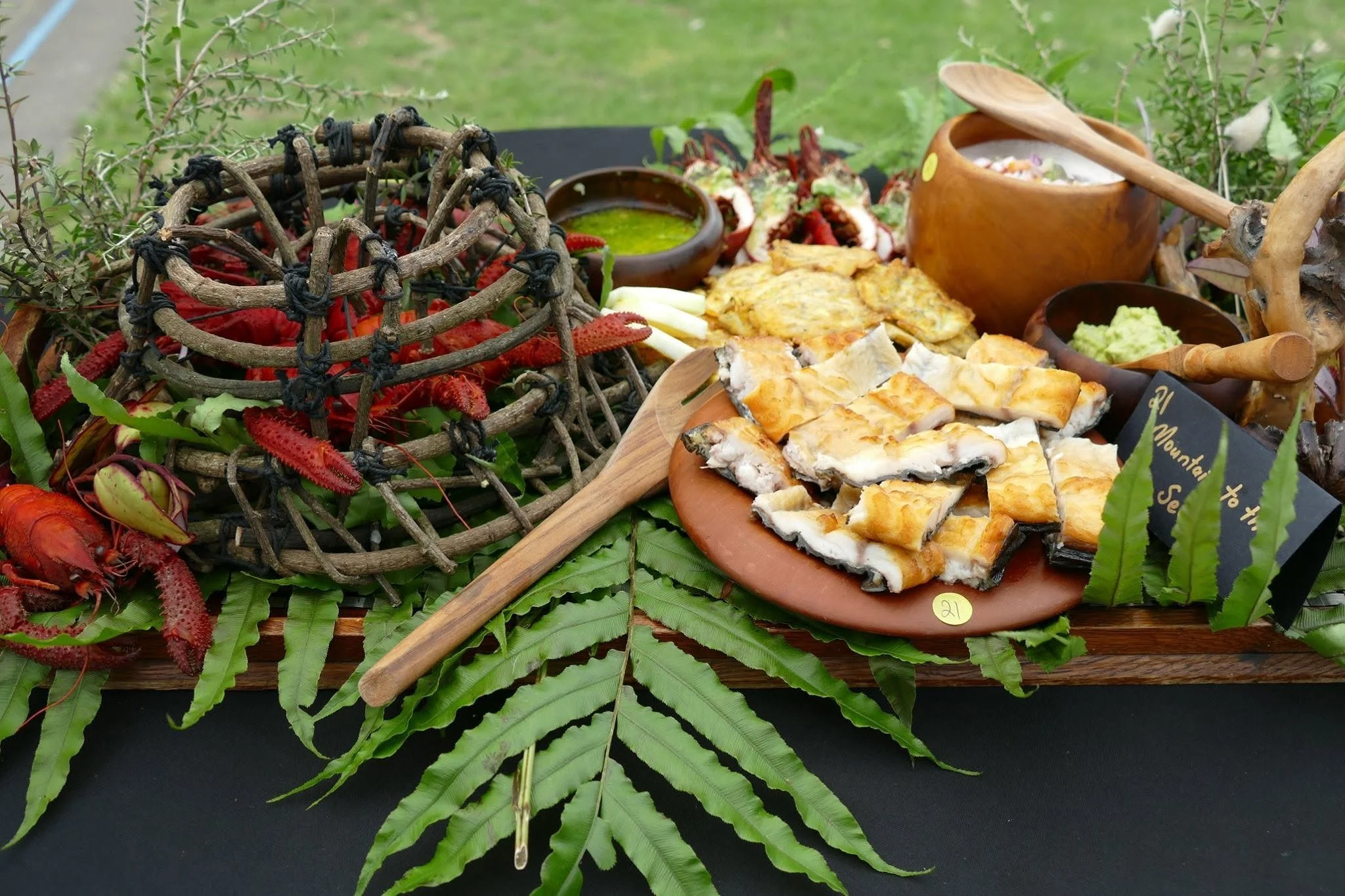 Display of traditional indigenous foods on a table outdoors, including grilled fish, a green sauce, guacamole, fresh herbs, and a decorative crown made of twigs with crawfish around it.