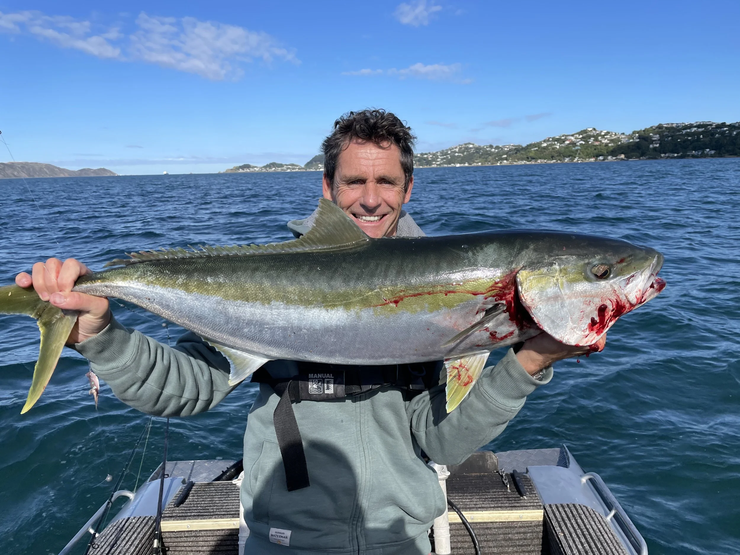 A man smiling on a boat holding a large fish with water and hills in the background.