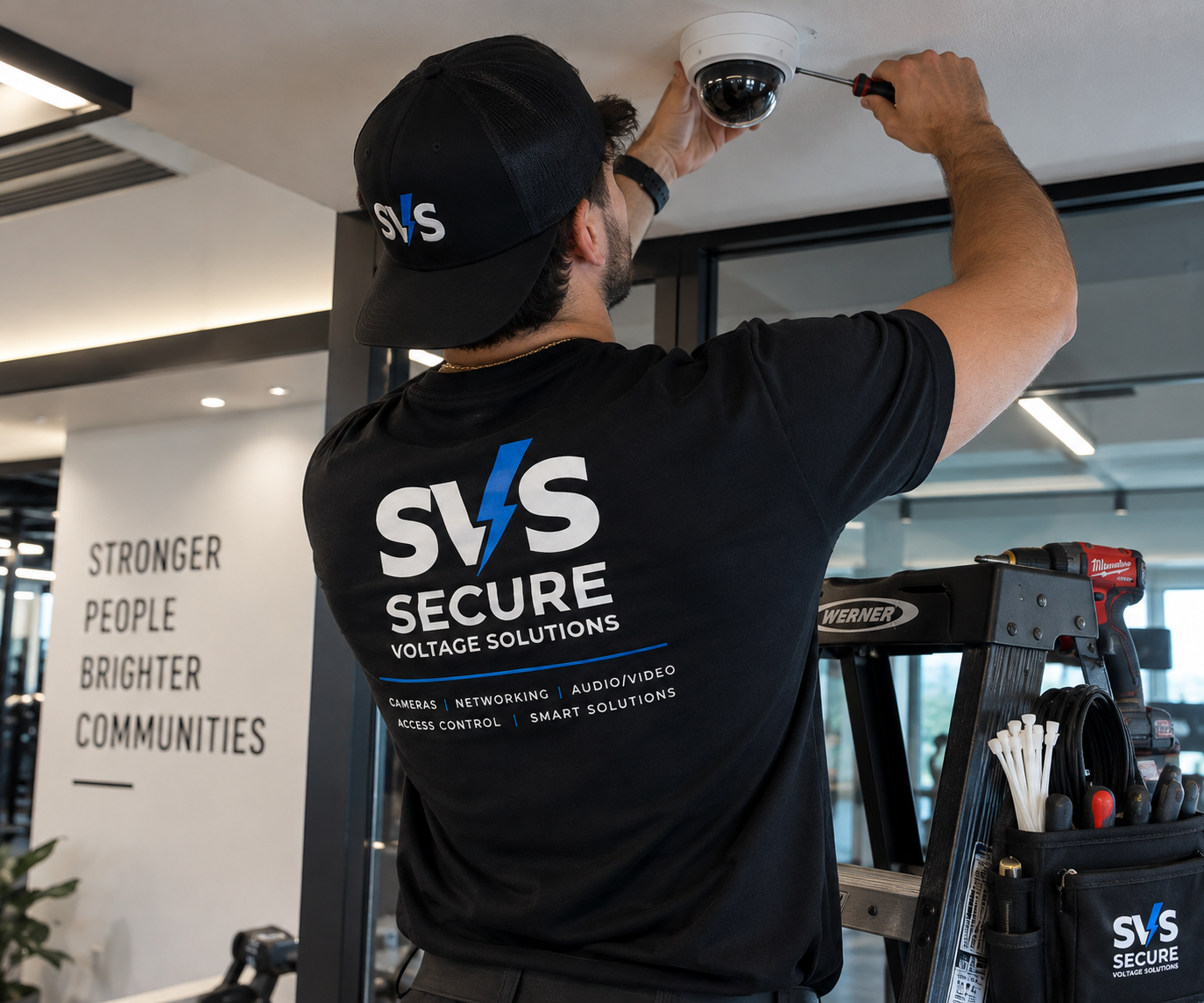 A technician wearing a black cap and t-shirt branded with "SVS Secure" is installing a security camera on the ceiling. There is a ladder and tools nearby, and the wall in the background has a sign with the words "Stronger People Brighter Communities."