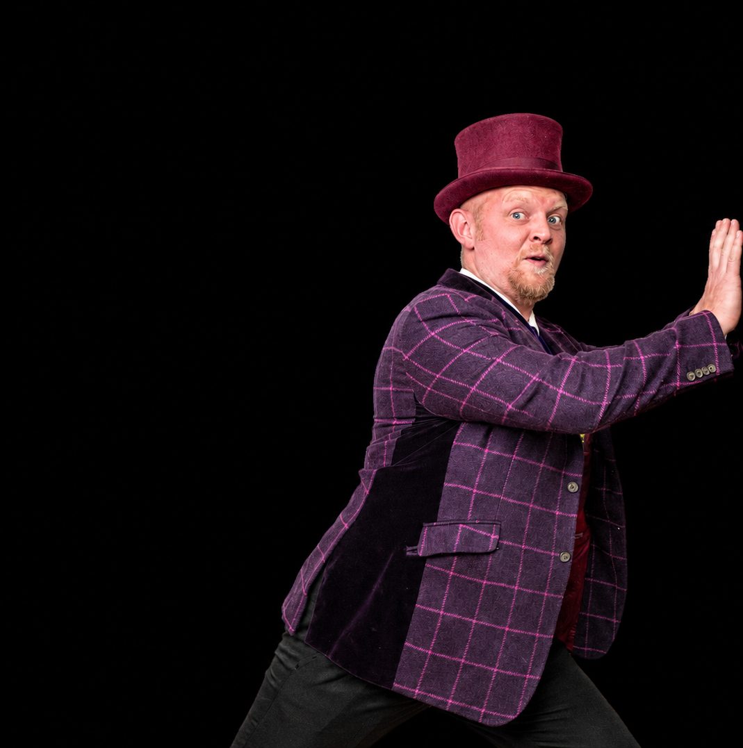 A man with red hair and beard wearing a purple plaid jacket and a maroon top hat, posing with one hand raised against a black background.